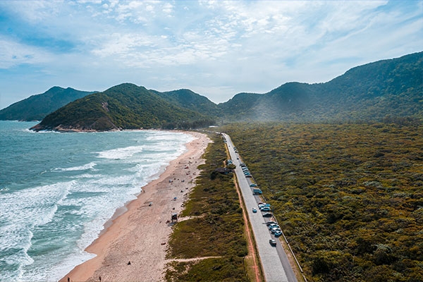 Foto aérea de Grumari, no Rio de Janeiro. É dia. Aparecem o mar, a areia, uma estrada ao lado da praia e uma mata com morros ao fundo