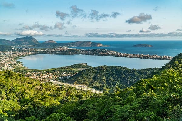 Foto da Baía de Piratinga, em Niterói. Aparecem o céu azul, a mata, residências e o mar