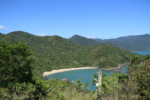 Foto da Praia do Secreto, no RIo de Janeiro. Ao fundo, aparecem um morro, o céu azul, o mar e árvores.