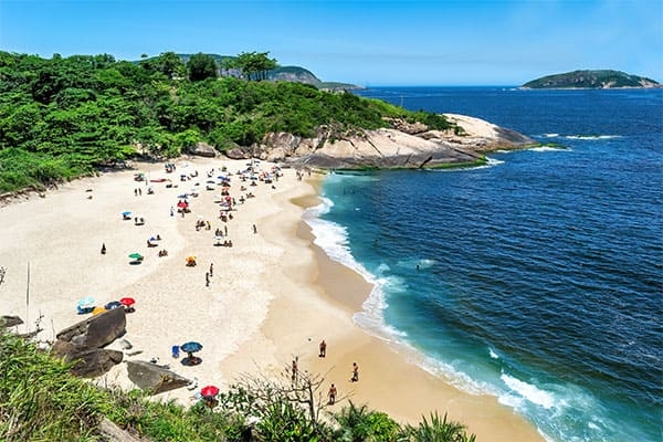 Foto da Praia do Sossego, em Niterói. Ao fundo, aparecem o céu azul, a areia com movimento de pessoas e o mar.