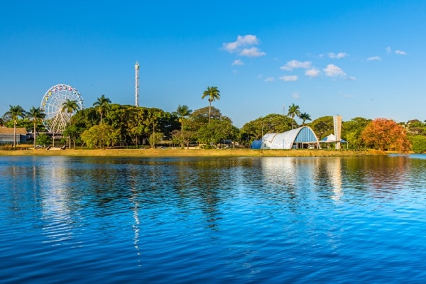 Vista da lagoa da Pampulha, em Belo Horizonte. É dia, com poucas nuvens no céu e a água tem um azul vibrante, onde é possível ver a igreja São Francisco ao fundo.