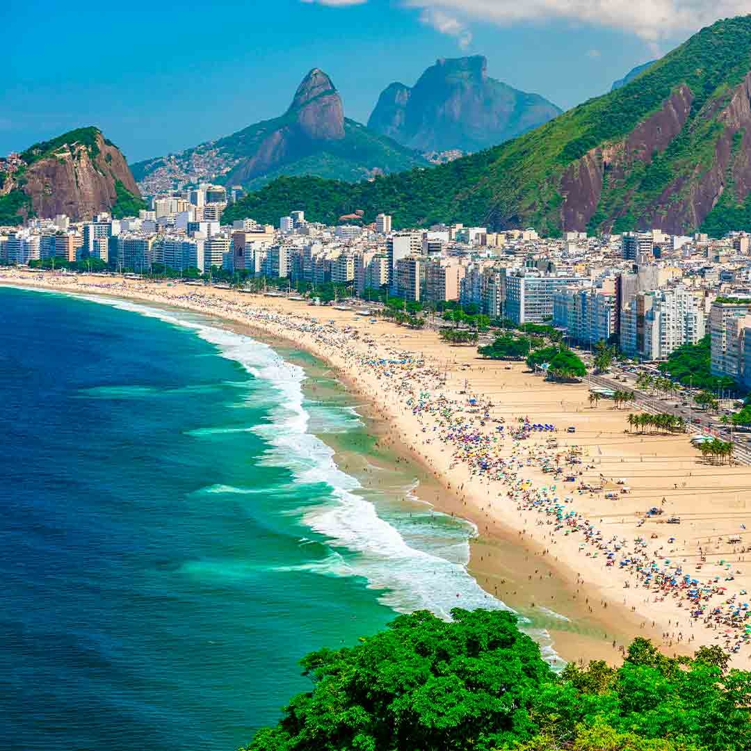 Foto da Praia de Copacabana, no Rio de Janeiro. Ao fundo, aparecem morros e o céu azul. À frente, o mar, a areia com diversas pessoas e guarda-sóis, além dos prédios na orla.