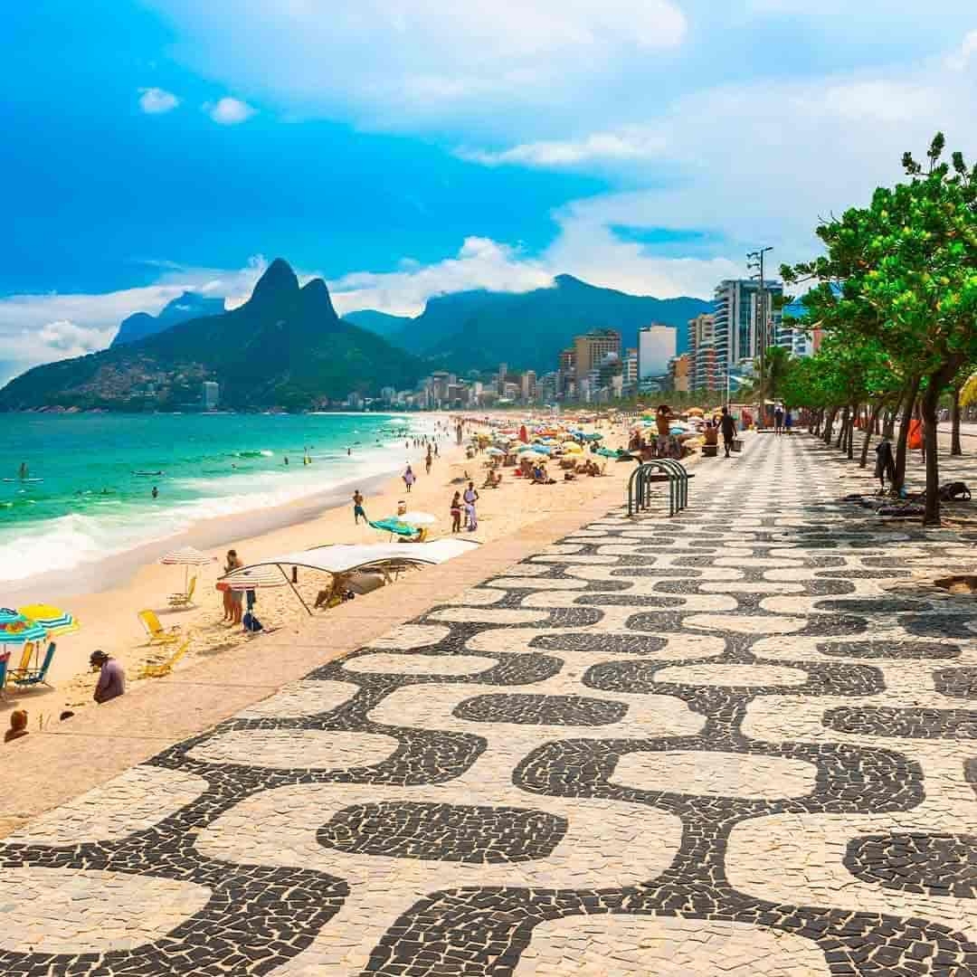 Foto da Praia de Ipanema, no Rio de Janeiro. Ao fundo, aparecem morros e o céu azul. À frente, o calçadão de Ipanema, o mar e diversas pessoas e guarda-sóis na areia.