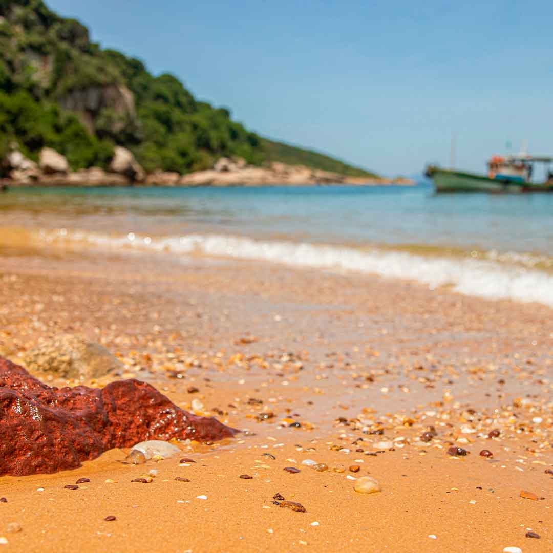 Foto do Arquipélago de Santana, em Macaé. Ao fundo, aparecem um morro, o céu azul, o mar e uma canoa. À frente, uma faixa de areia com muitas conchas e pedras pequenas.