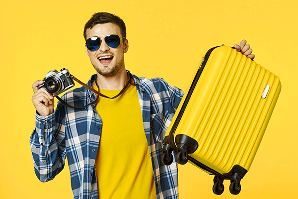 Imagem mostra um homem de óculos escuros, segurando em uma das mãos uma câmera e na outra mão uma mala de mão amarela