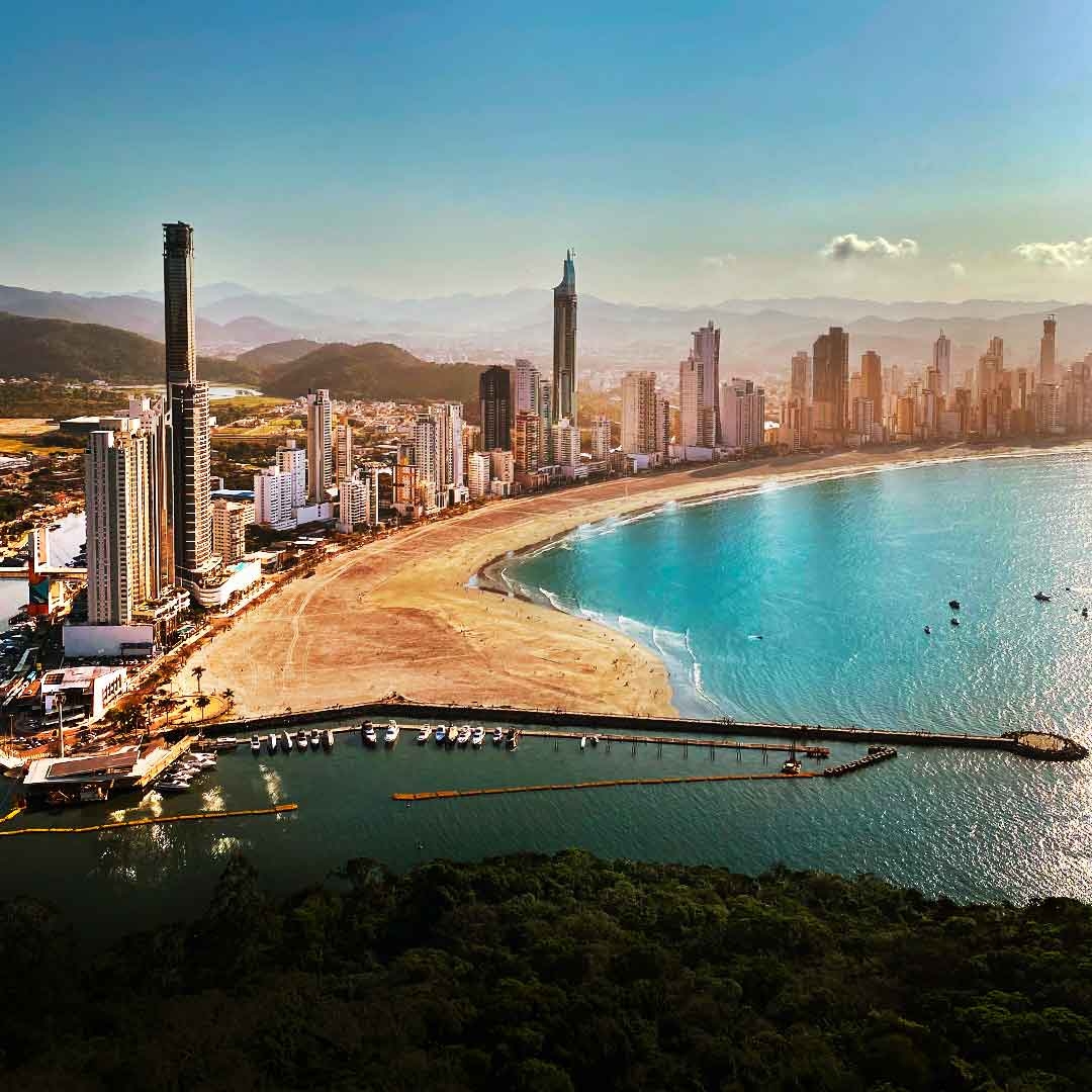 Foto da Praia Central, em Balneário Camboriú. Aparecem o céu azul, prédios e arranha-céus, o mar, e a areia.