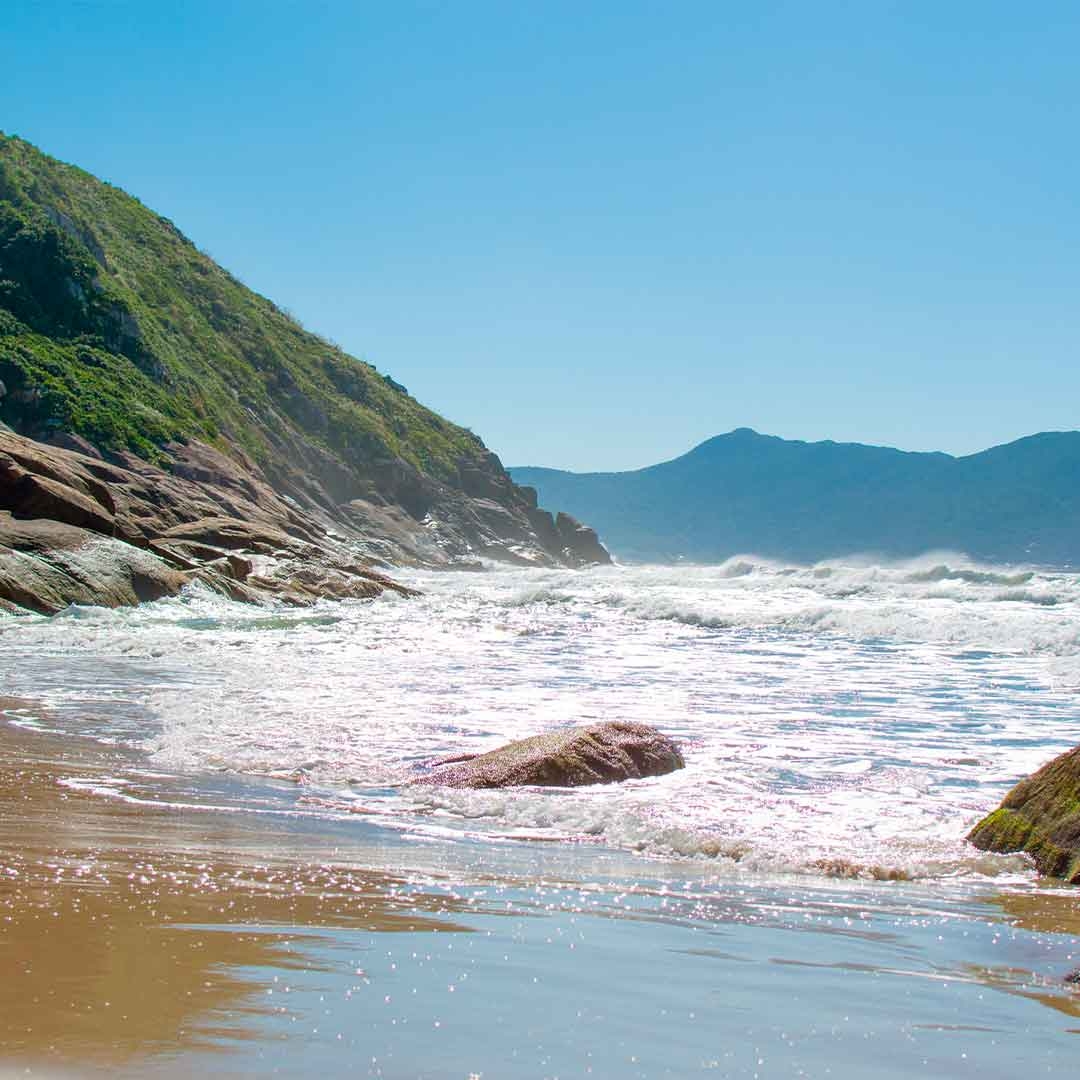 Foto da Praia da Solidão, em Florianópolis. Ao fundo, aparecem morros e o céu azul. À frente, o mar e a areia.