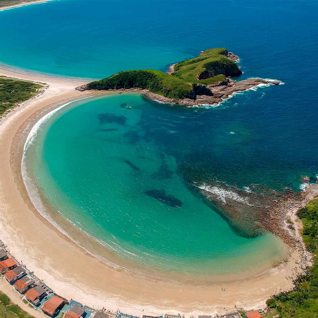 Foto aérea da Praia das Conchas, em Cabo Frio. É dia. Aparecem o mar, a areia, cabanas e uma ilha.