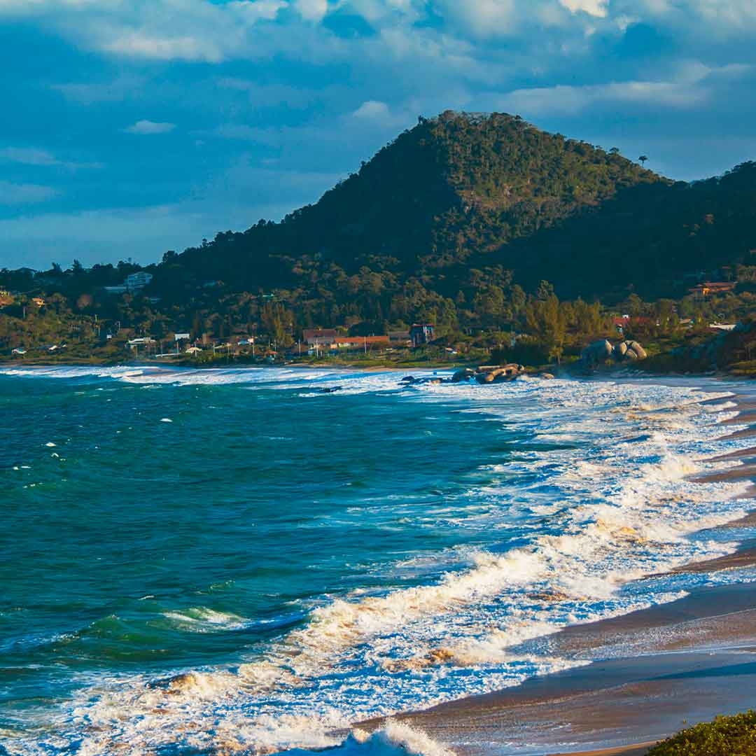 Foto da Praia do Estaleirinho, em Florianópolis. Ao fundo, aparecem morros, o céu azul e algumas residências. À frente, o mar.