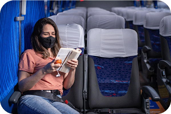Imagem ilustra uma mulher de máscara lendo revista sentada dentro de um ônibus.